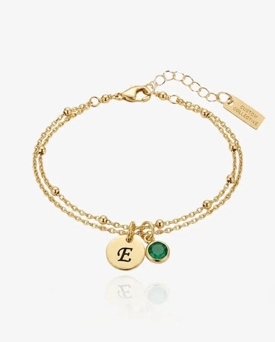 Gold bracelet with a round disc engraved with the initial E and a green birthstone charm, on a double chain, shown flat